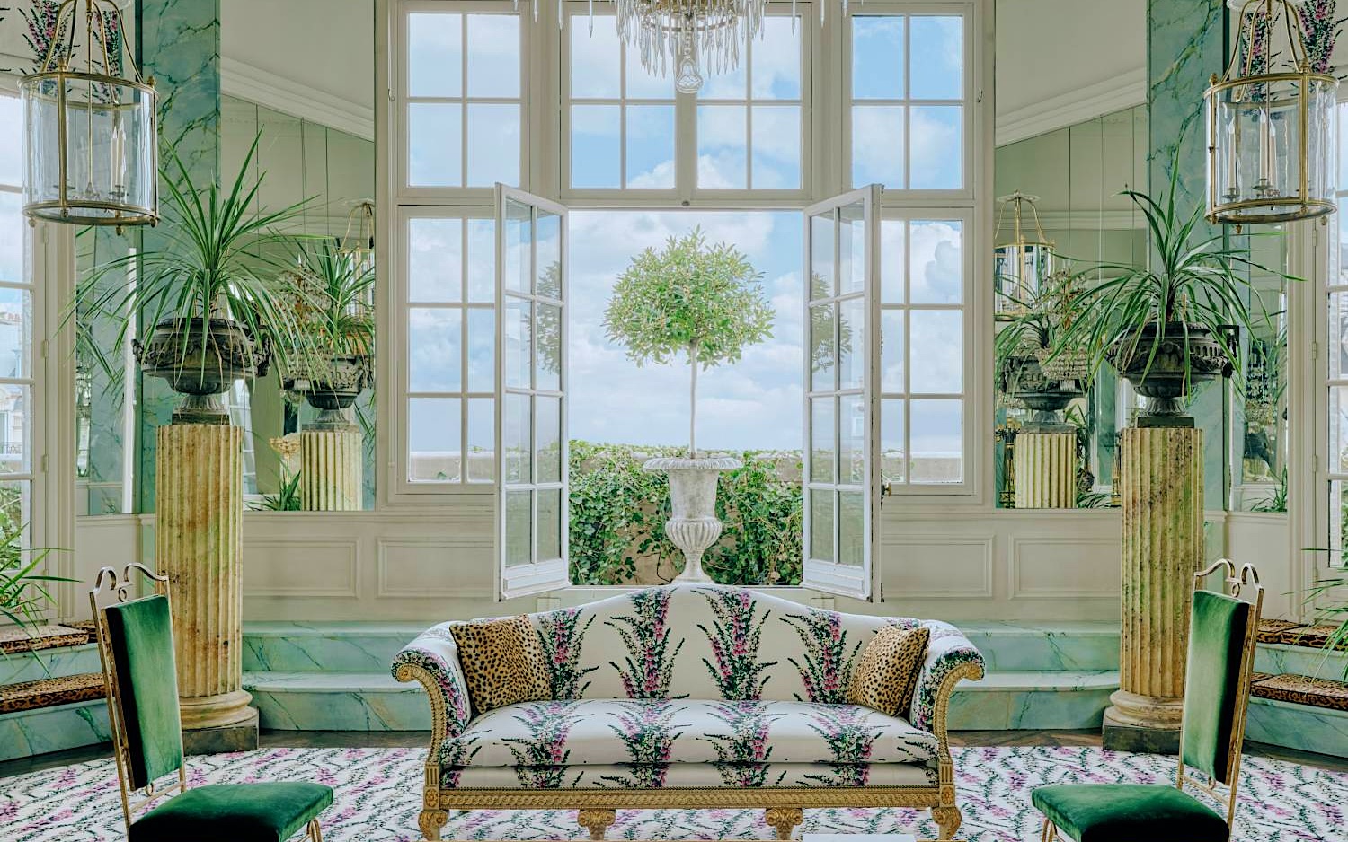 The salon at Le Style Beaujard: a crystal chandelier above a floral sofa with leopard cushions, two green velvet chairs, marble columns with palms, and open French windows onto a clipped bay tree